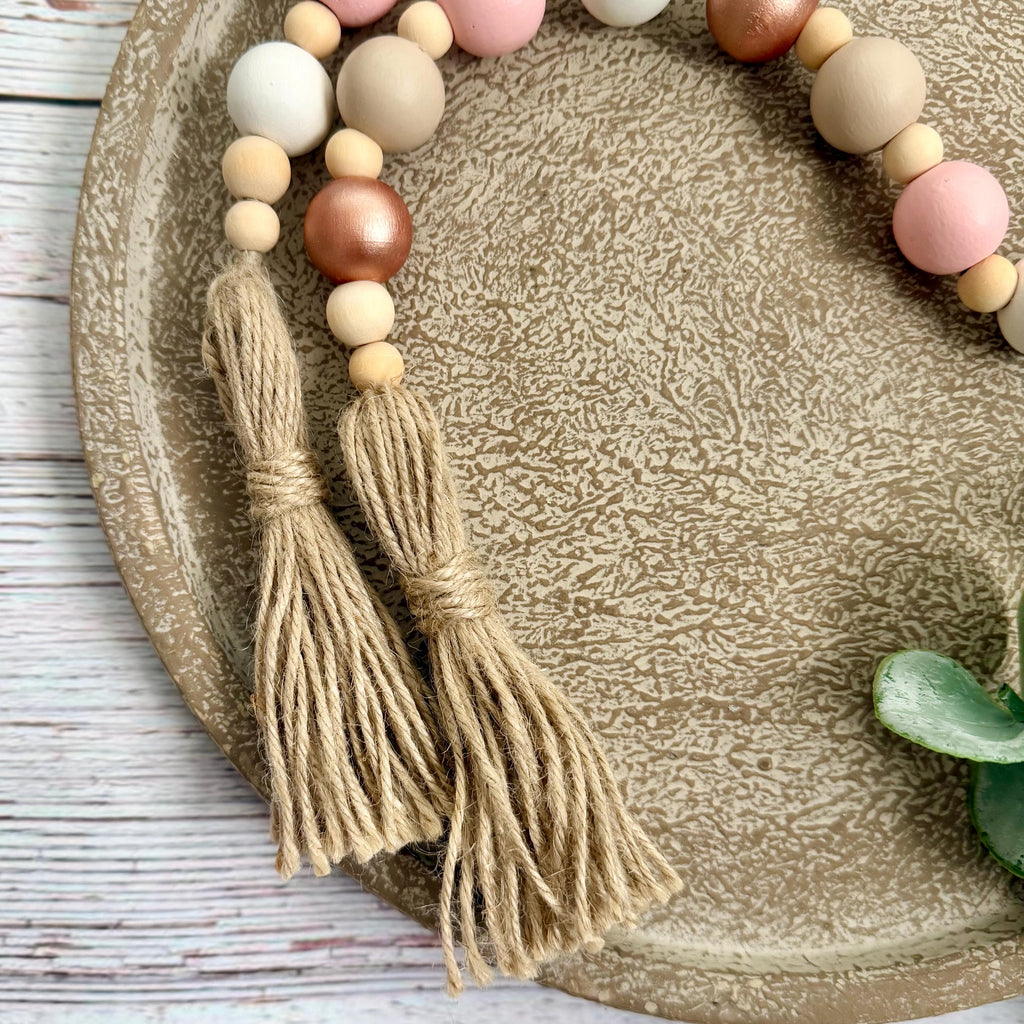 A decorative beaded garland with two large jute tassels, arranged on a textured beige ceramic plate over a whitewashed wood surface. The beads include natural wood, blush pink, white, and metallic rose gold.