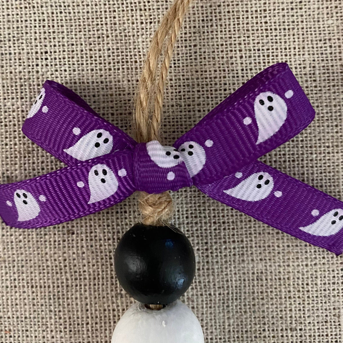 A Halloween hanging ornament featuring a purple grosgrain ribbon bow printed with white ghosts, suspended from a natural jute twine string with a black wooden bead and white stone bead below, set against a beige burlap background.