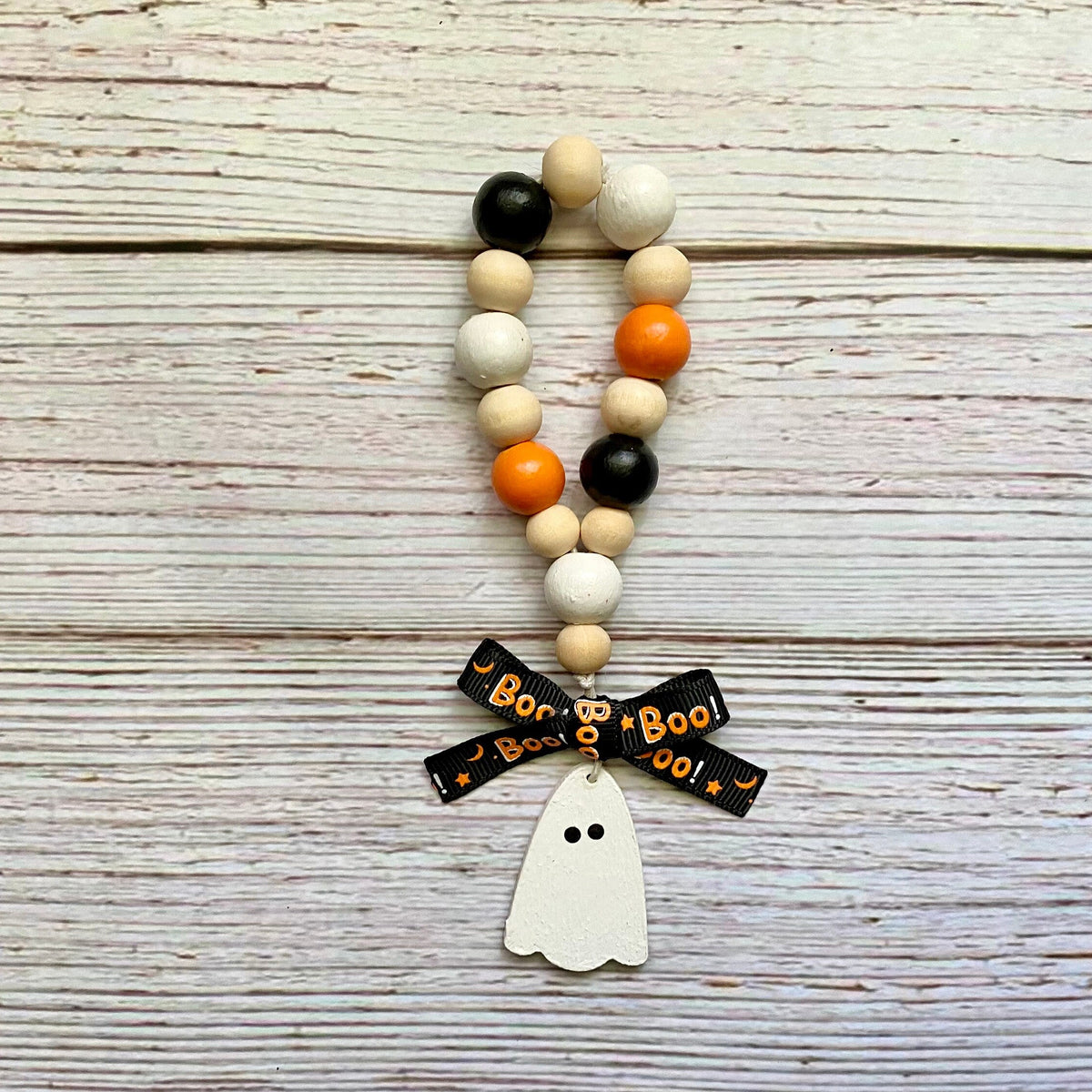 A beaded loop with natural wood beads, orange and black beads, a black ribbon bow printed with 'Boo! Boo! Boo!', and a white ghost charm, displayed on a rustic white-washed wooden background.