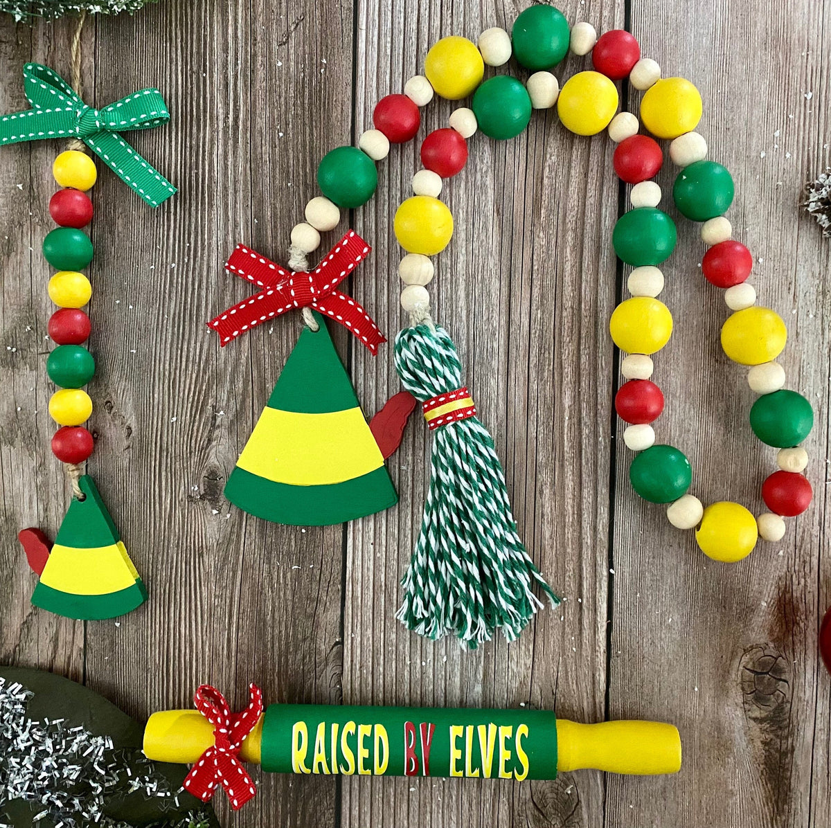 A festive Christmas bead garland set displayed on a rustic wooden background. The set includes a long beaded garland with red, green, yellow, and white beads, a green and yellow wooden elf hat ornament, a green and yellow wooden Christmas tree ornament, a green and white striped tassel, and a green and yellow rolling pin with 'RAISED BY ELVES' written on it.