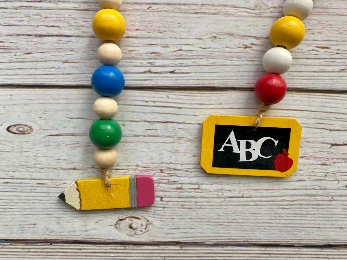 Two colorful school-themed bead garlands displayed on a whitewashed wooden background. One garland features yellow, cream, blue, green, and pink beads ending in a wooden pencil charm.