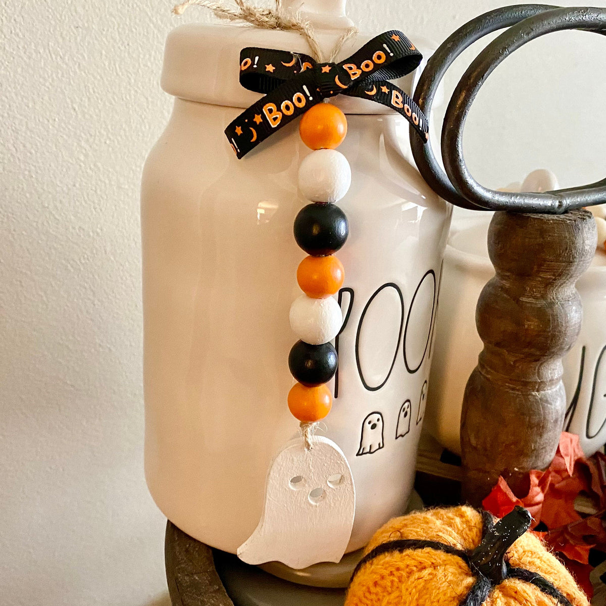 A ceramic Halloween-themed jar with a white lid and black 'boo' lettering, decorated with a black ribbon, orange and white beads, and a white ghost ornament. Other seasonal decor items, including a small knitted pumpkin and autumn leaves, are arranged around it on a tabletop against a plain wall.