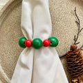 A close-up of a beaded ring-style accessory with alternating green and red round beads, wrapped around a white napkin on a textured beige surface with a pinecone prop nearby.