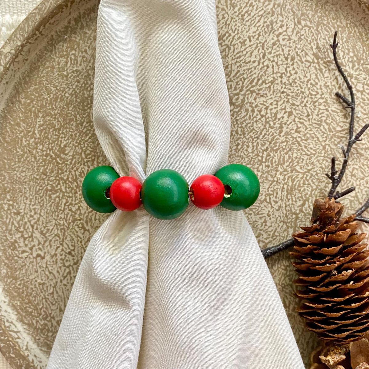 A close-up of a beaded ring-style accessory with alternating green and red round beads, wrapped around a white napkin on a textured beige surface with a pinecone prop nearby.