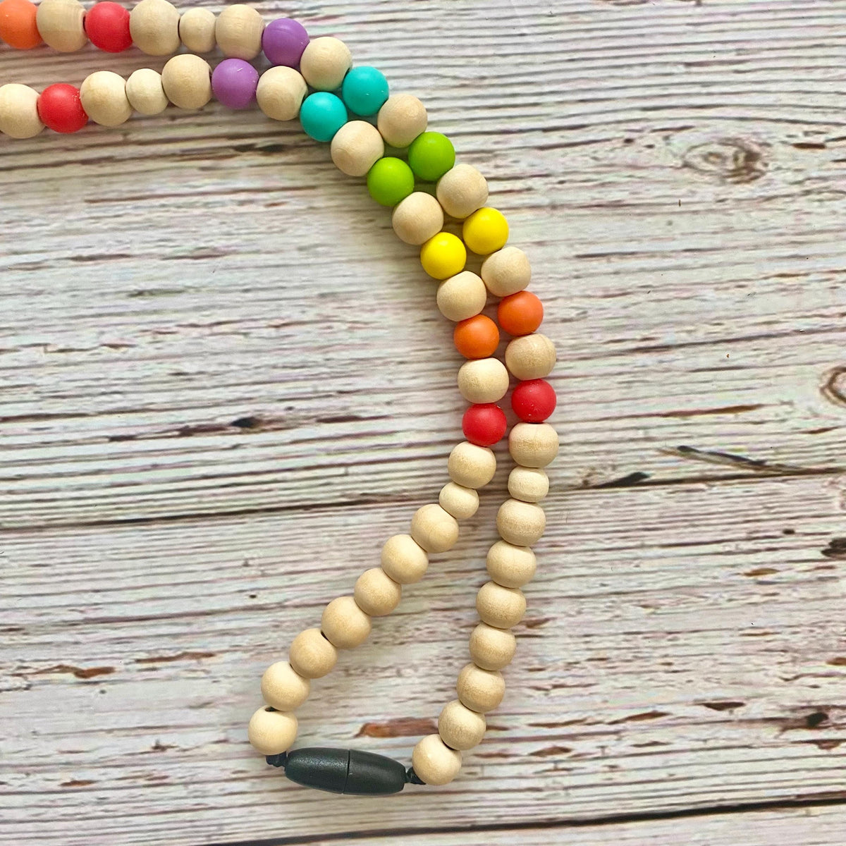 A wooden bead necklace or rosary laid on a weathered white wood surface, featuring natural light wood beads with a rainbow sequence of colored beads including red, orange, yellow, green, turquoise, and purple, finished with a black toggle clasp.
