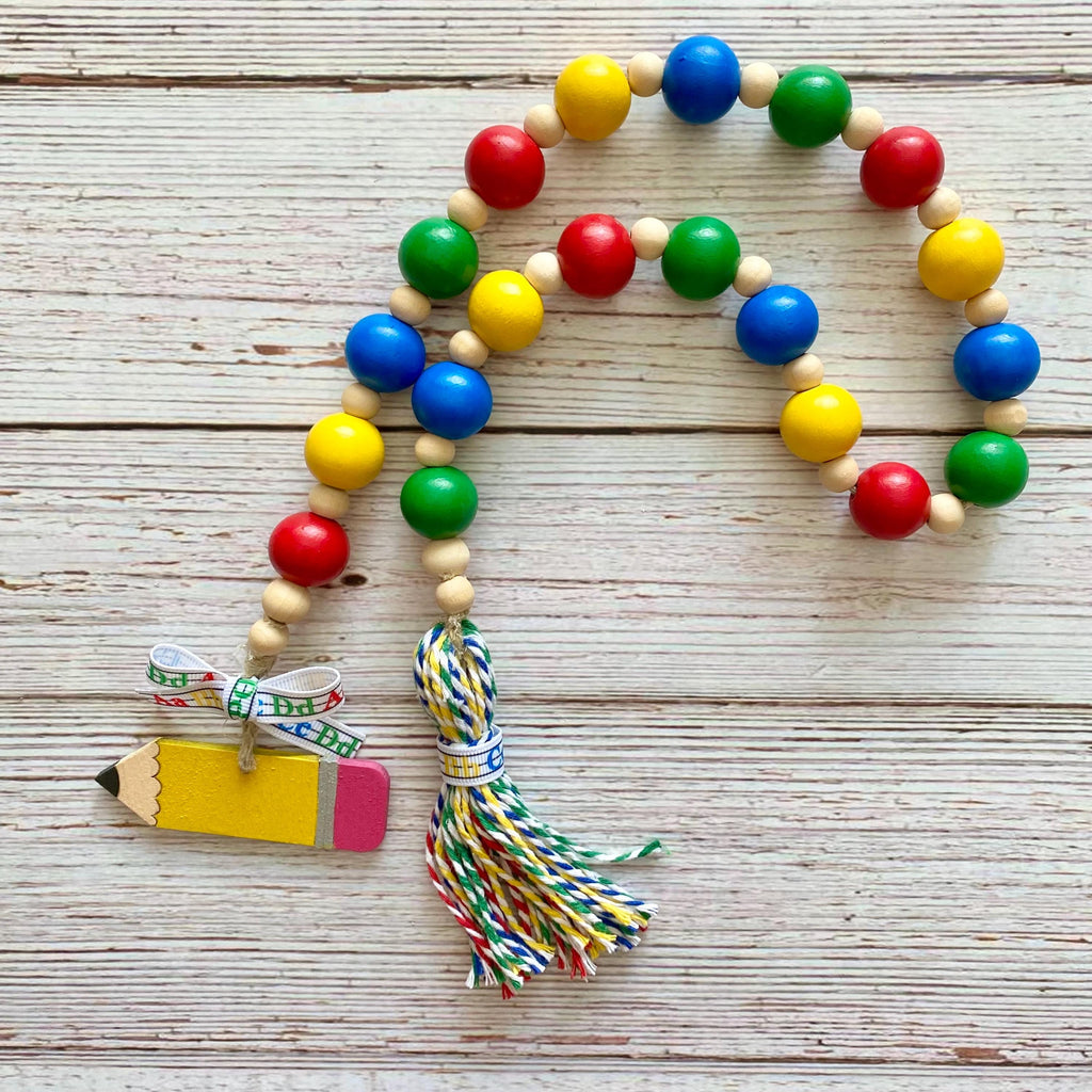 A colorful wooden bead garland with red, yellow, green, and blue beads, a tassel, and a pencil-shaped charm, displayed on a light wood plank background.