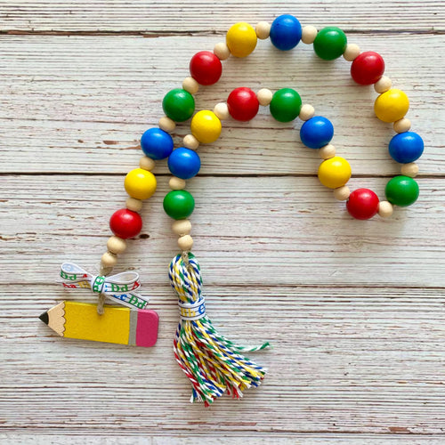A colorful wooden bead garland with red, yellow, green, and blue beads, a tassel, and a pencil-shaped charm, displayed on a light wood plank background.