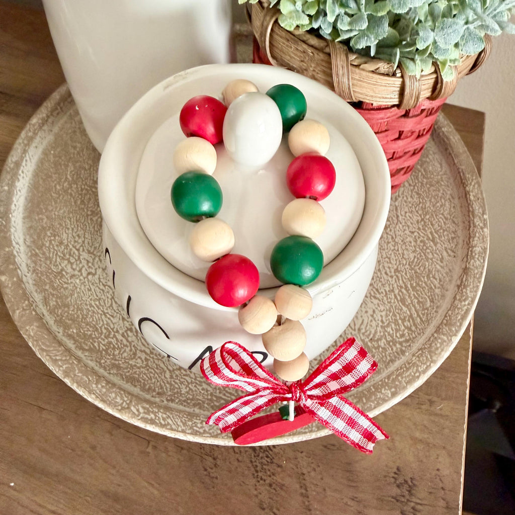 A red and green-themed wooden bead garland with red, green, and natural wood beads, tied with a red and white gingham ribbon and a red wooden apple, displayed on a white ceramic container.