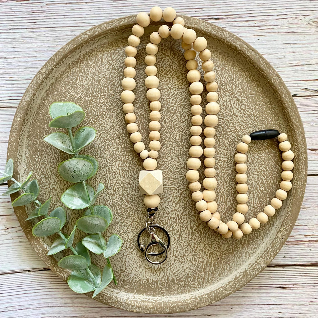 A beaded necklace made of light natural wood beads, styled on a round textured tray with green eucalyptus leaves. The necklace features a geometric wooden charm and a metal keychain pendant.