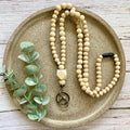 A beaded necklace made of light natural wood beads, styled on a round textured tray with green eucalyptus leaves. The necklace features a geometric wooden charm and a metal keychain pendant.