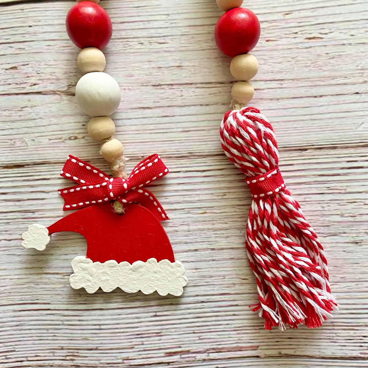 A handmade Christmas bead garland featuring red and natural wood beads, a red Santa hat ornament with a white-trimmed border and red ribbon bow, and a red-and-white striped cotton tassel, displayed on a light wood plank surface.