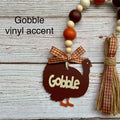 A fall-themed turkey decoration with a dark brown wooden turkey body, beige lettering that says 'Gobble,' a red-and-beige gingham ribbon bow, and a matching tassel. The item is shown on a whitewashed wood background with a beaded garland above it and the text 'Gobble vinyl accent' overlaid.