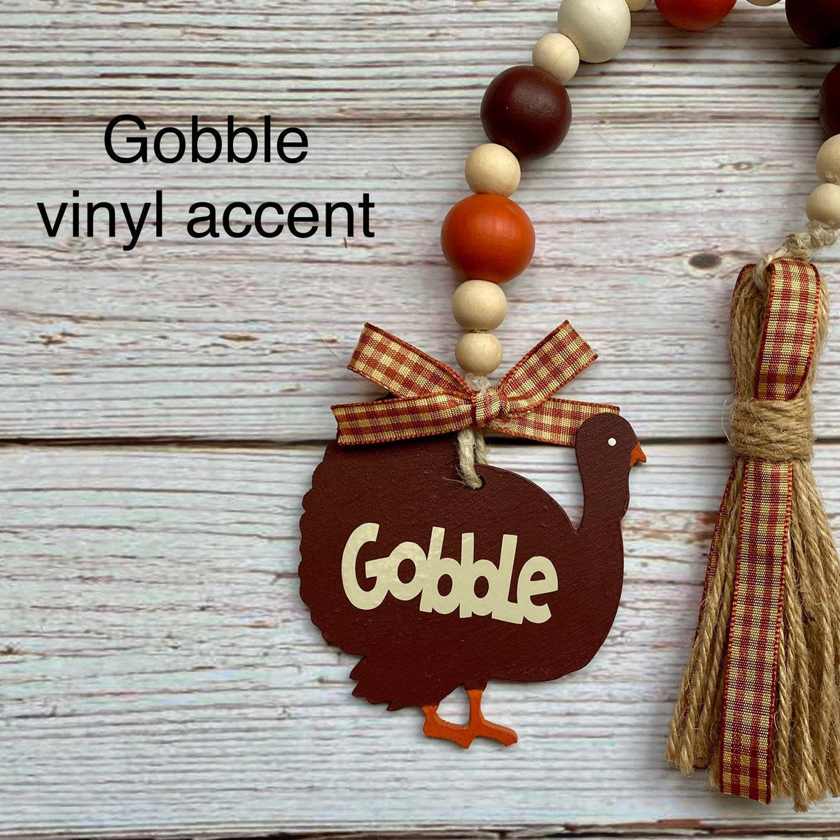 A fall-themed turkey decoration with a dark brown wooden turkey body, beige lettering that says 'Gobble,' a red-and-beige gingham ribbon bow, and a matching tassel. The item is shown on a whitewashed wood background with a beaded garland above it and the text 'Gobble vinyl accent' overlaid.