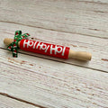 A wooden rolling pin with a bright red painted barrel, white 'Ho Ho Ho!' lettering, and a green holiday-themed ribbon bow, displayed on a rustic white-washed wooden surface.