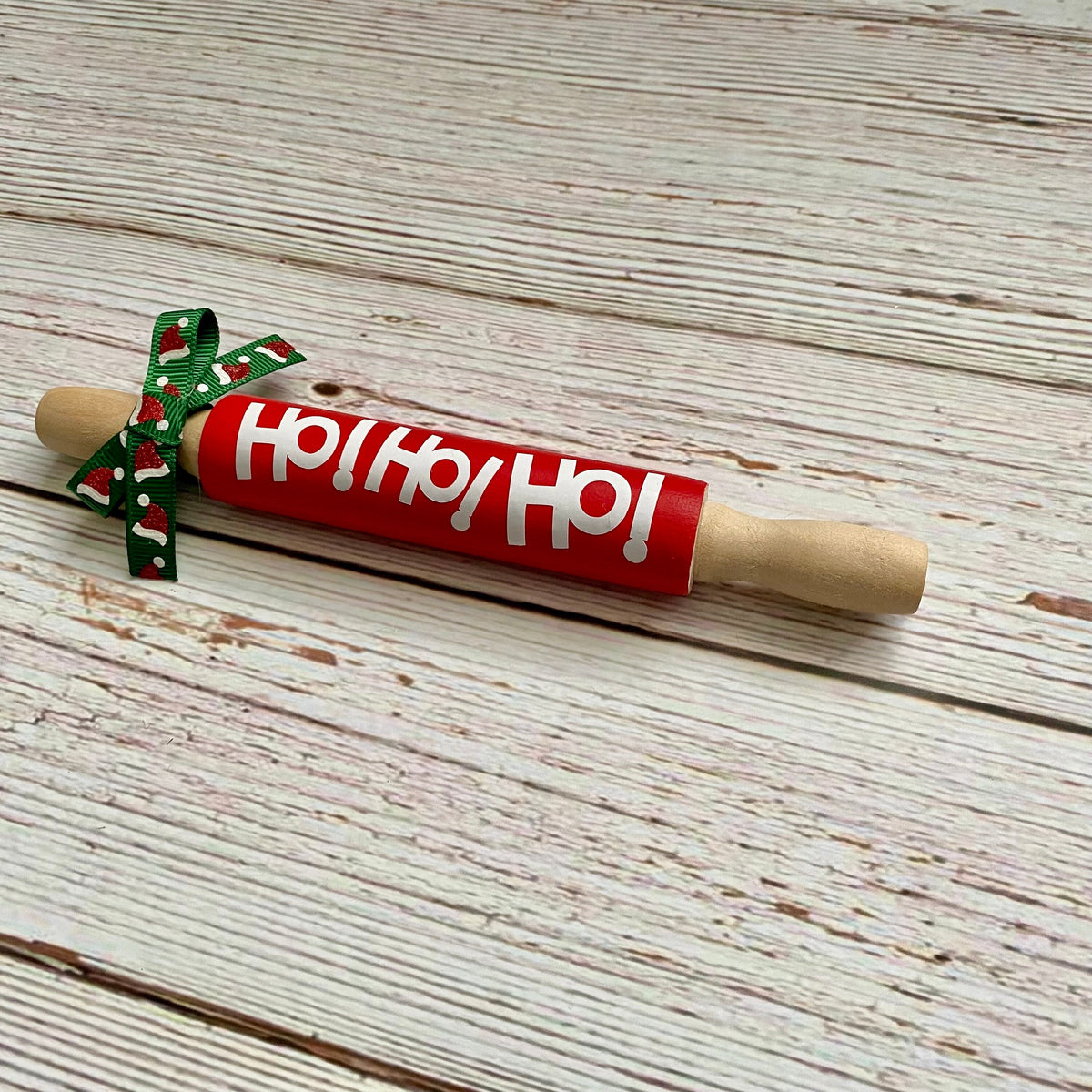 A wooden rolling pin with a bright red painted barrel, white 'Ho Ho Ho!' lettering, and a green holiday-themed ribbon bow, displayed on a rustic white-washed wooden surface.
