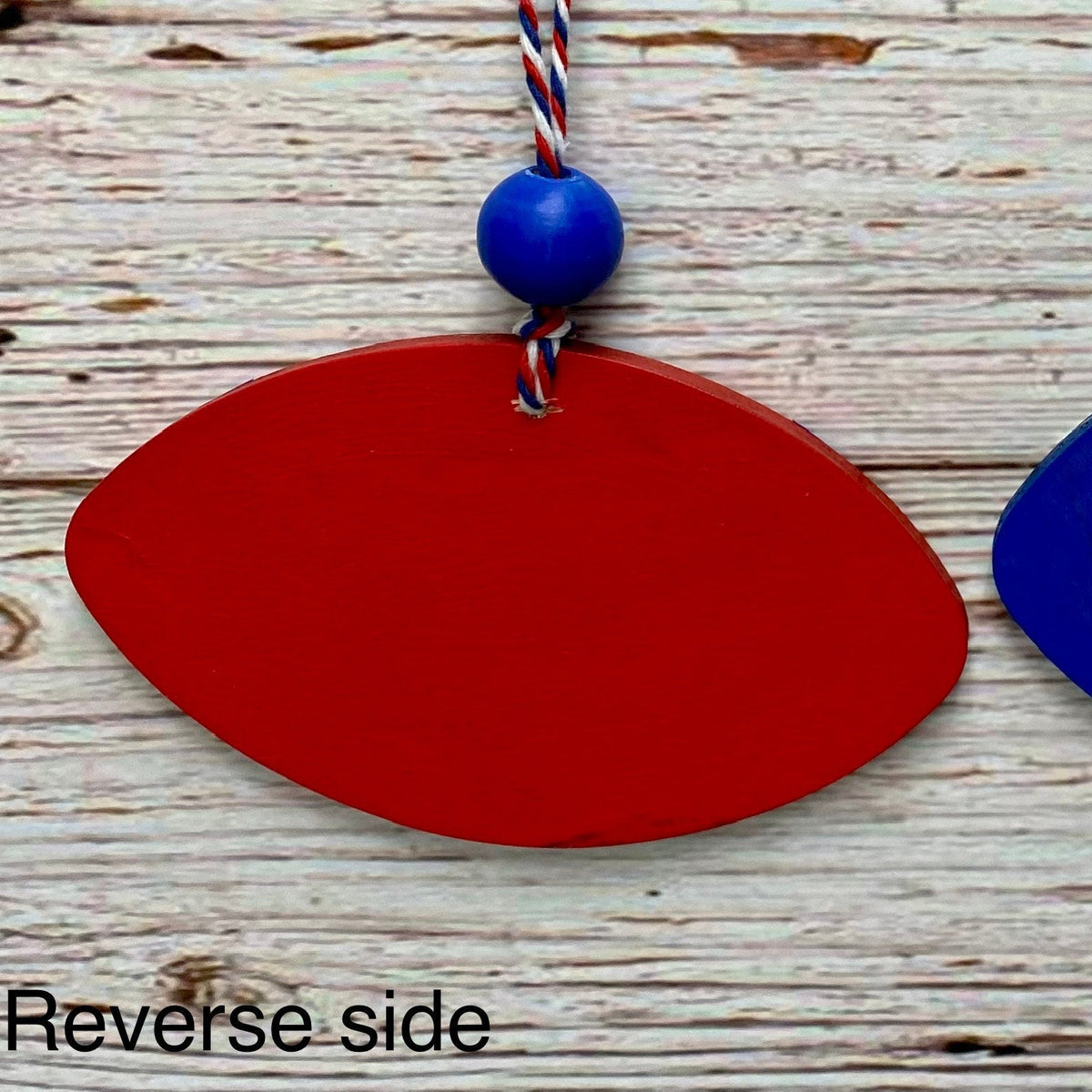 A red wooden ornament or plaque with a blue bead and red, white, and blue string, shown on a weathered white wood background with the text 'Reverse side' at the bottom.