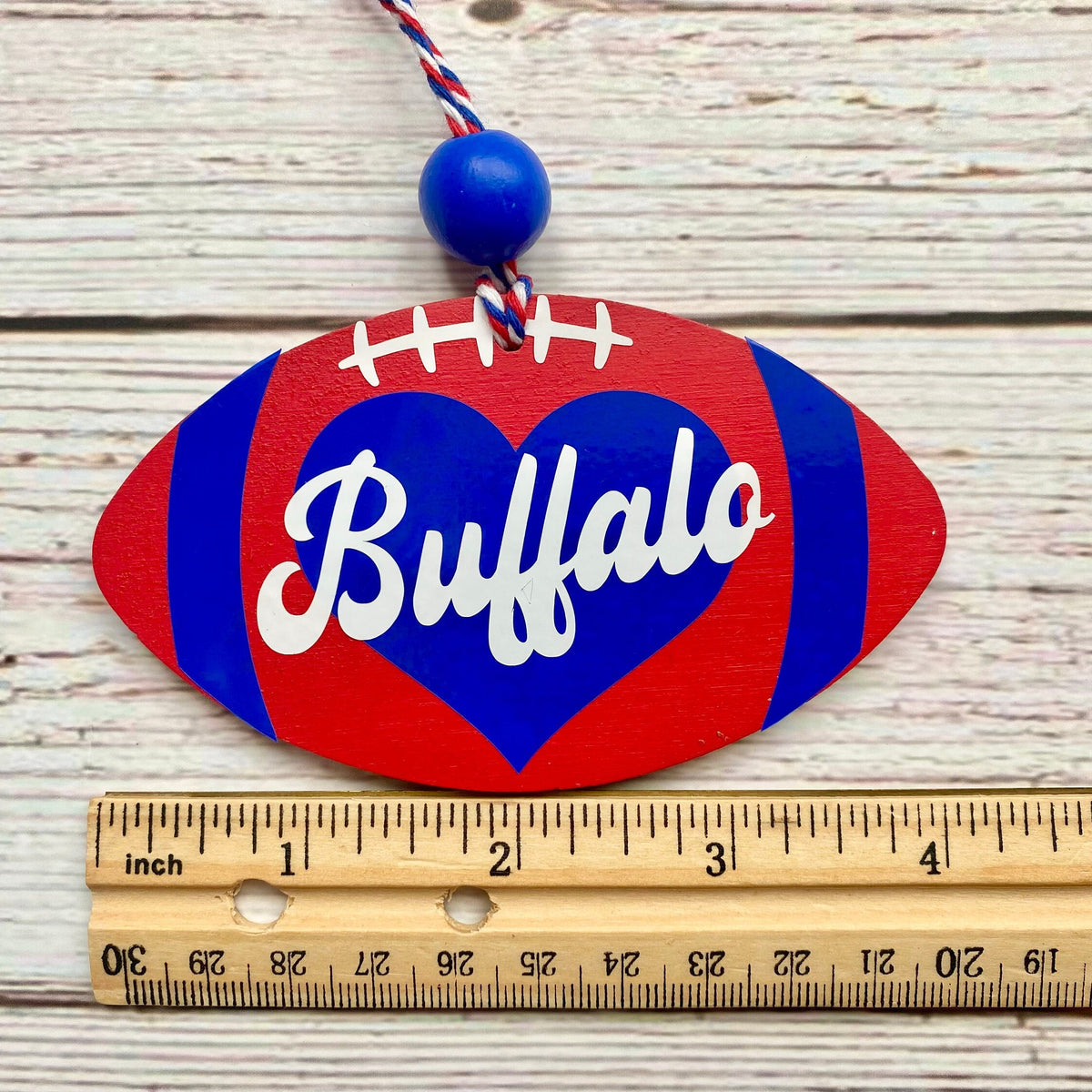 A wooden football-shaped ornament with a red and blue buffalo heart design and the word 'Buffalo' in white script, shown on a whitewashed wood background with a ruler for scale.