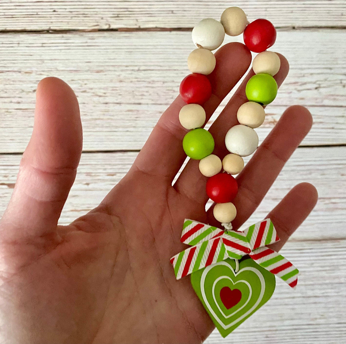 A hand holding a beaded Christmas ornament or keychain charm with red, green, and cream wooden beads and a green heart-shaped hanging tag with a red heart cutout and a red-and-green striped ribbon bow, against a whitewashed wood plank background.