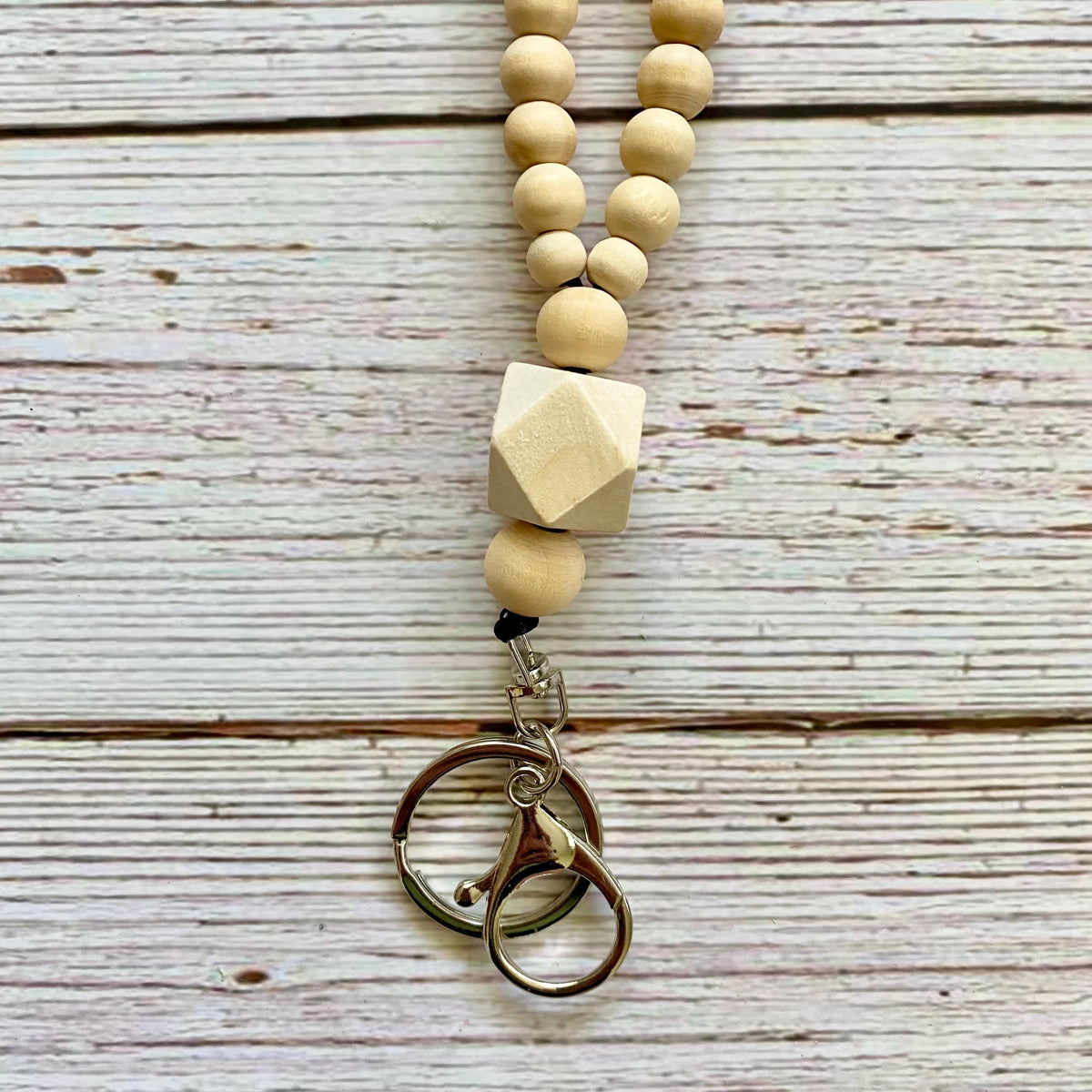A beaded necklace or keychain charm made of natural light wood beads and a faceted wooden cube, with a silver-tone metal keyring clasp, laid flat on a whitewashed wooden surface.