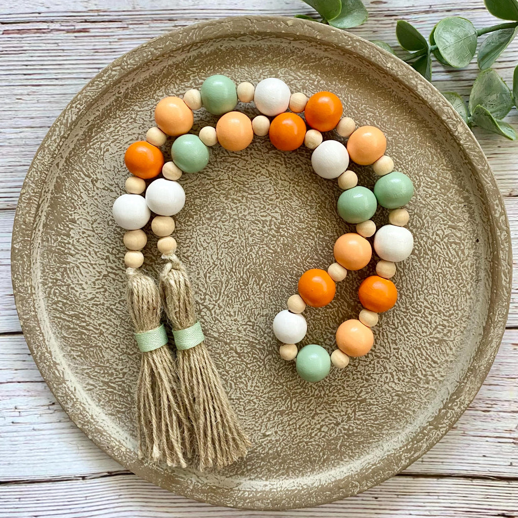 A beaded tassel garland with orange, white, and sage green wooden beads and two beige tassels, displayed on a round textured ceramic plate over a white wood background.