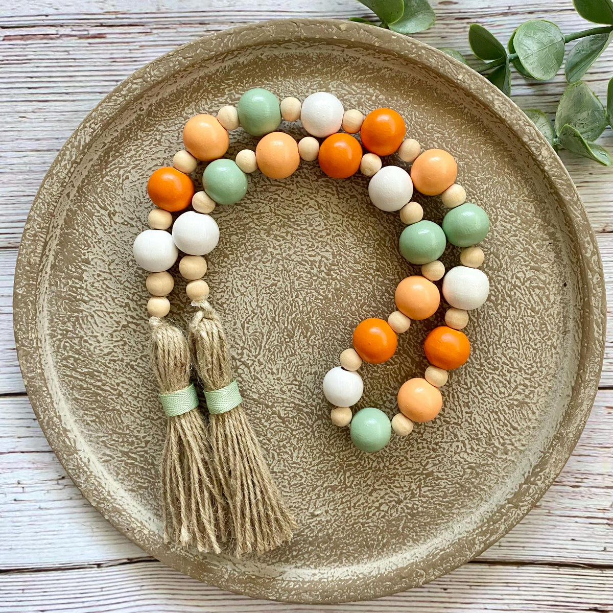 A beaded tassel garland with orange, white, and sage green wooden beads and two beige tassels, displayed on a round textured ceramic plate over a white wood background.