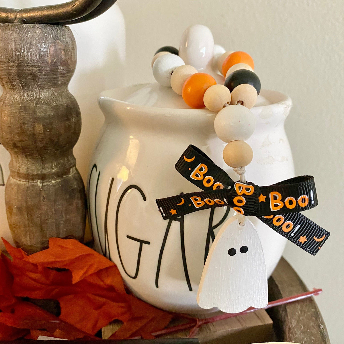 A white ceramic Halloween-themed jar or candle holder with black lettering, decorated with a beaded garland and a black ribbon bow printed with 'Boo!'.