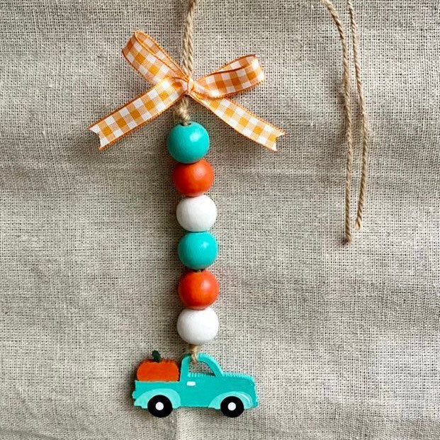 A handmade autumn beaded ornament hanging from a twine string, featuring a gingham orange and white ribbon bow at the top, alternating teal, orange, and white wooden beads, and a small teal pickup truck with an orange pumpkin at the bottom, displayed against a textured beige linen background.