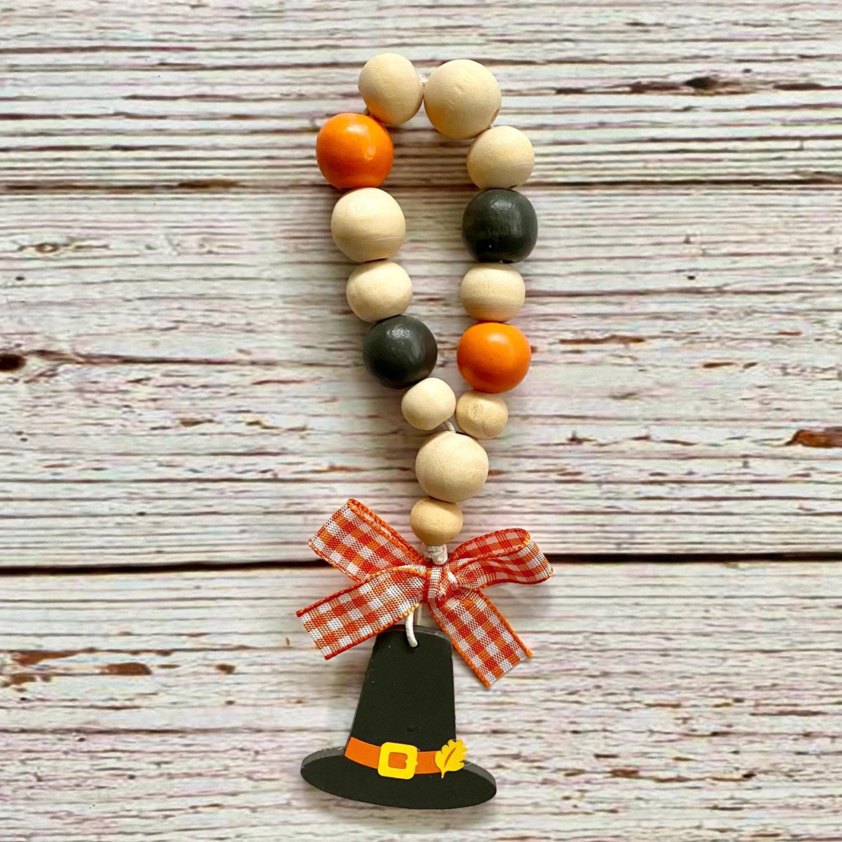 A wooden bead garland with orange, black, and natural beige beads, finished with a red and white gingham bow and a painted black pilgrim hat ornament, displayed on a rustic whitewashed wood background.
