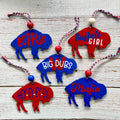 Six buffalo-shaped hanging ornaments in red and blue are arranged on a whitewashed wooden surface. Each ornament has a different phrase printed on it: BUFFALO, Buffalo GIRL, BIG DUBS, HELL-Y-EY-EY, and Mafia.