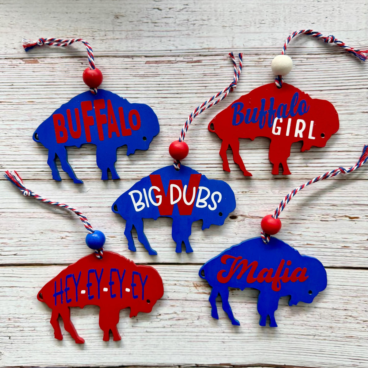 Six buffalo-shaped hanging ornaments in red and blue are arranged on a whitewashed wooden surface. Each ornament has a different phrase printed on it: BUFFALO, Buffalo GIRL, BIG DUBS, HELL-Y-EY-EY, and Mafia.