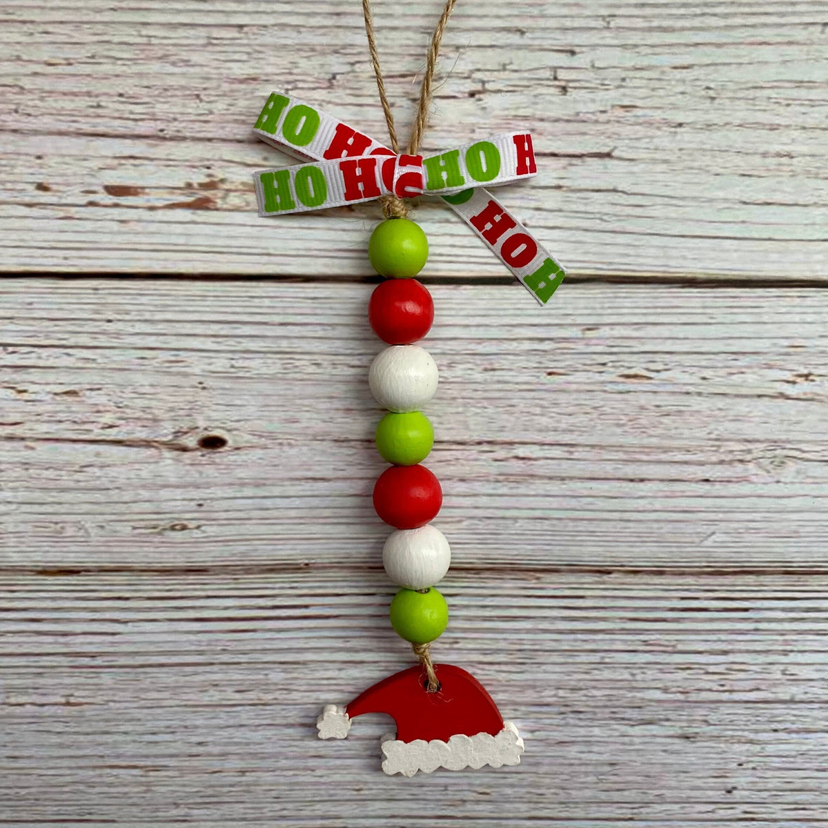 A Christmas hanging ornament made from wooden beads in red, green, and white, with a Santa hat charm and a bow made from HO Ho HO pieces, hanging against a rustic white-washed wood background.