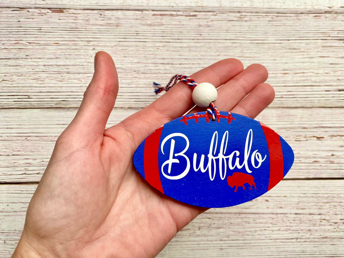 A hand holding a custom wooden football-shaped ornament painted blue and red with the word 'Buffalo' in white script and a small red buffalo silhouette. The ornament has a red, white, and blue string loop with a white bead, set against a light wood plank background.