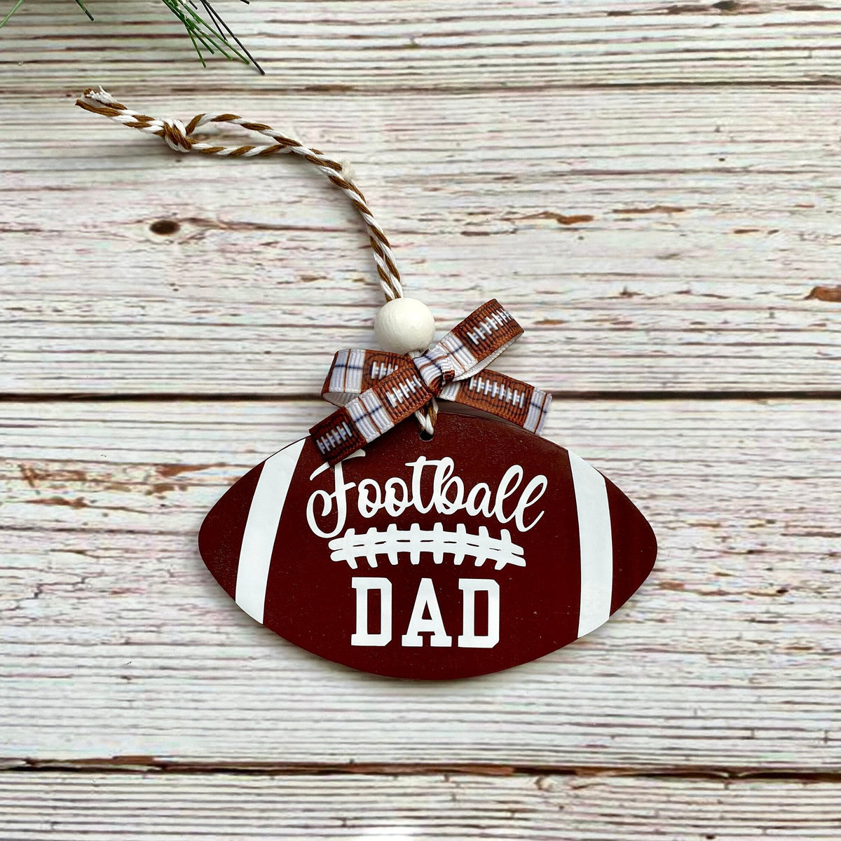 A football-shaped wooden ornament hanging on a rustic whitewashed wood surface. The ornament is dark red with white stripes, white 'Football DAD' lettering, and a small plaid ribbon bow with a white bead and twine loop.