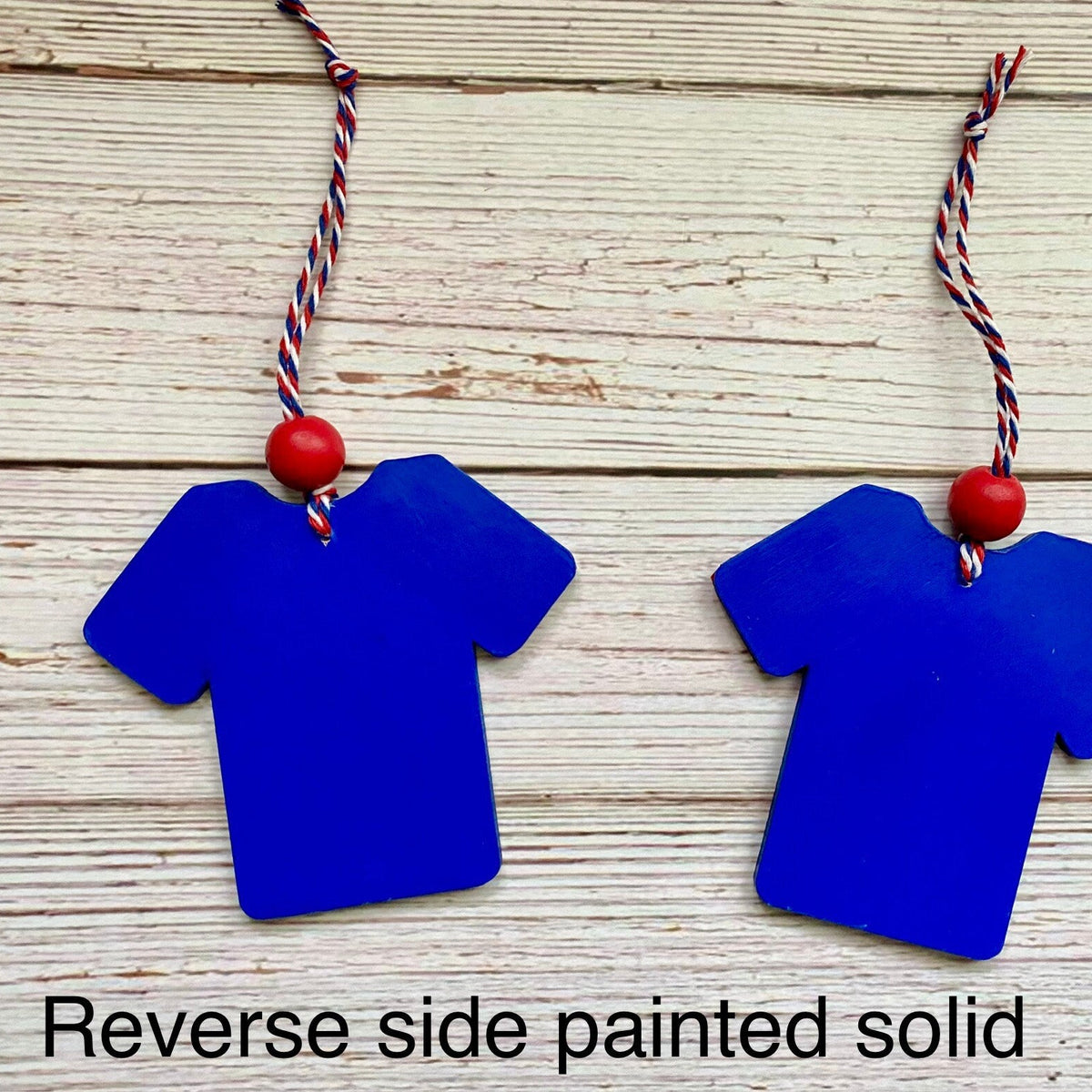 Two blue t-shirt-shaped ornaments hanging on a light wood background, each with a red bead and red, white, and blue string. Text at the bottom reads 'Reverse side painted solid'.