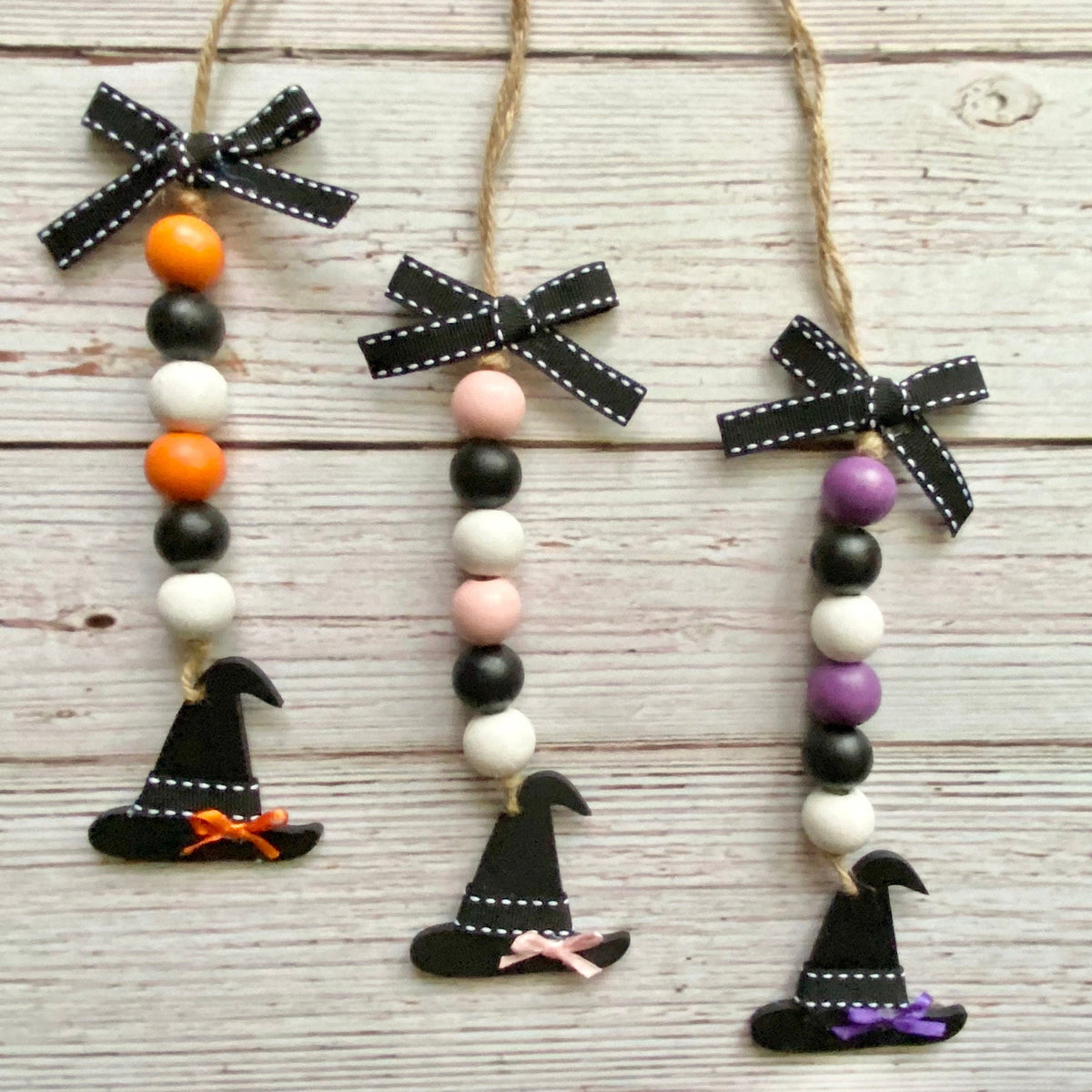 Three Halloween bead garlands with black ribbon bows, black, white, orange, pink, and purple beads, and black witch hats hanging on a whitewashed wooden background.