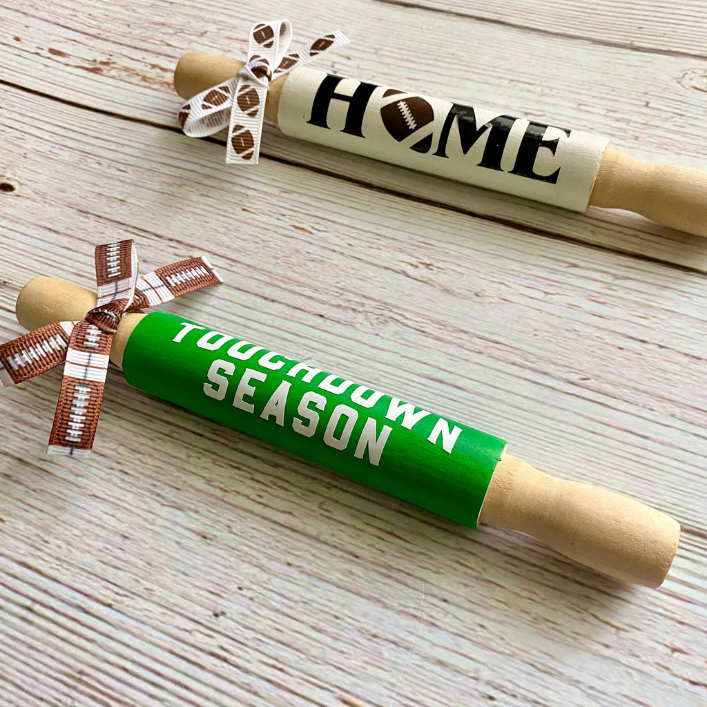 Two wooden rolling pins displayed on a whitewashed wood background. One rolling pin is cream-colored with 'HOME' written in black and a football-shaped 'O', tied with a brown and white football-patterned ribbon.