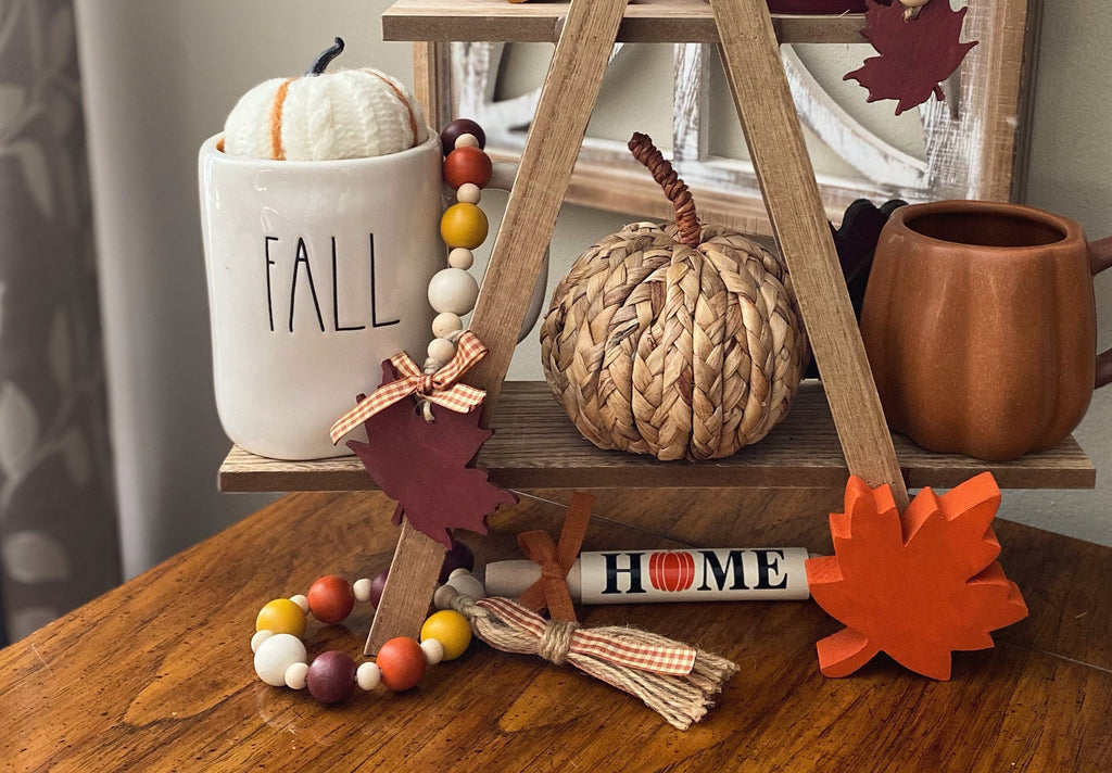 A styled autumn tabletop scene with a wooden A-frame shelf holding a woven pumpkin, a white ceramic mug with the word FALL, a terracotta cup, colorful bead garlands, orange and burgundy paper leaves, and a small white letter scroll spelling HOME.