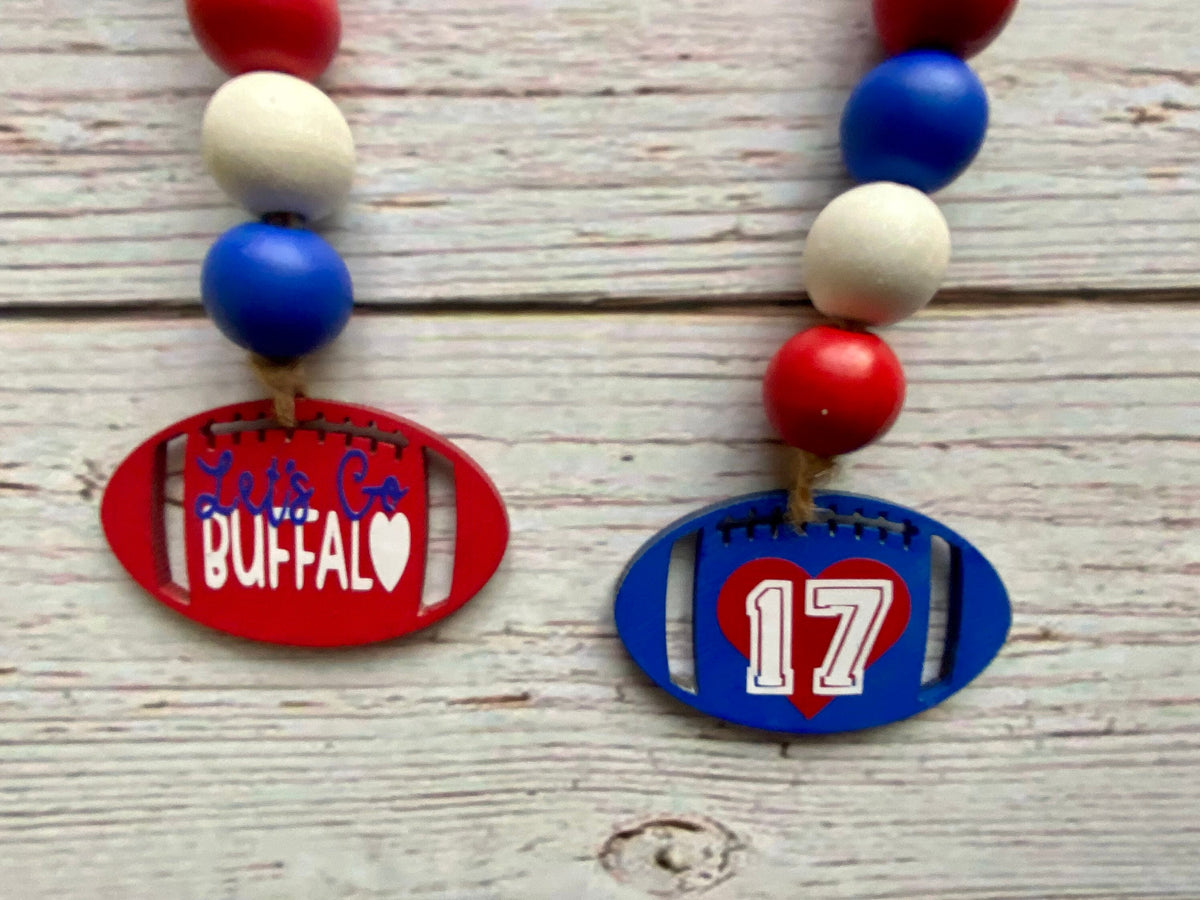 A beaded garland laid flat on a whitewashed wood surface, featuring red, white, and blue wooden beads with football-shaped end pieces. One red piece reads 'Let's Go BUFFALO' in white script with a heart, and the blue piece shows the number 17 over a red heart.