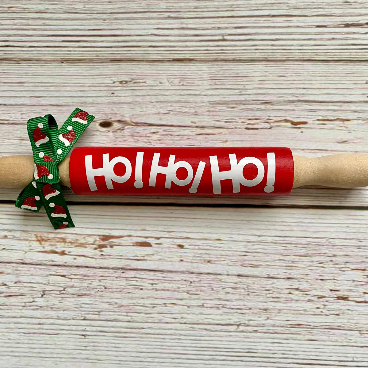 A wooden rolling pin with a bright red center section printed with 'Ho! Ho! Ho!' in white, plus a green holiday-themed ribbon bow with red and white accents, placed on a whitewashed wood plank surface.