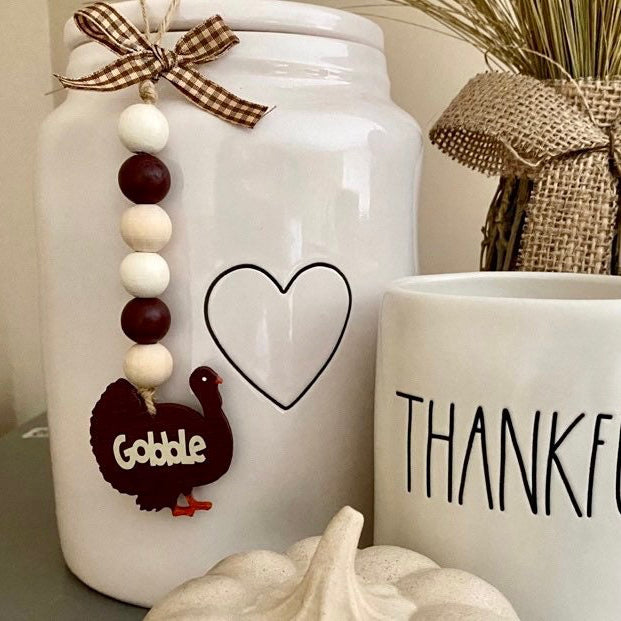 A white ceramic mason jar decorated with a brown gingham ribbon, wooden beads, and a turkey ornament that reads 'Gobble,' next to a white mug with 'THANKFUL' printed in black. The scene includes a burlap-wrapped decorative bundle and a small pumpkin on a tabletop.