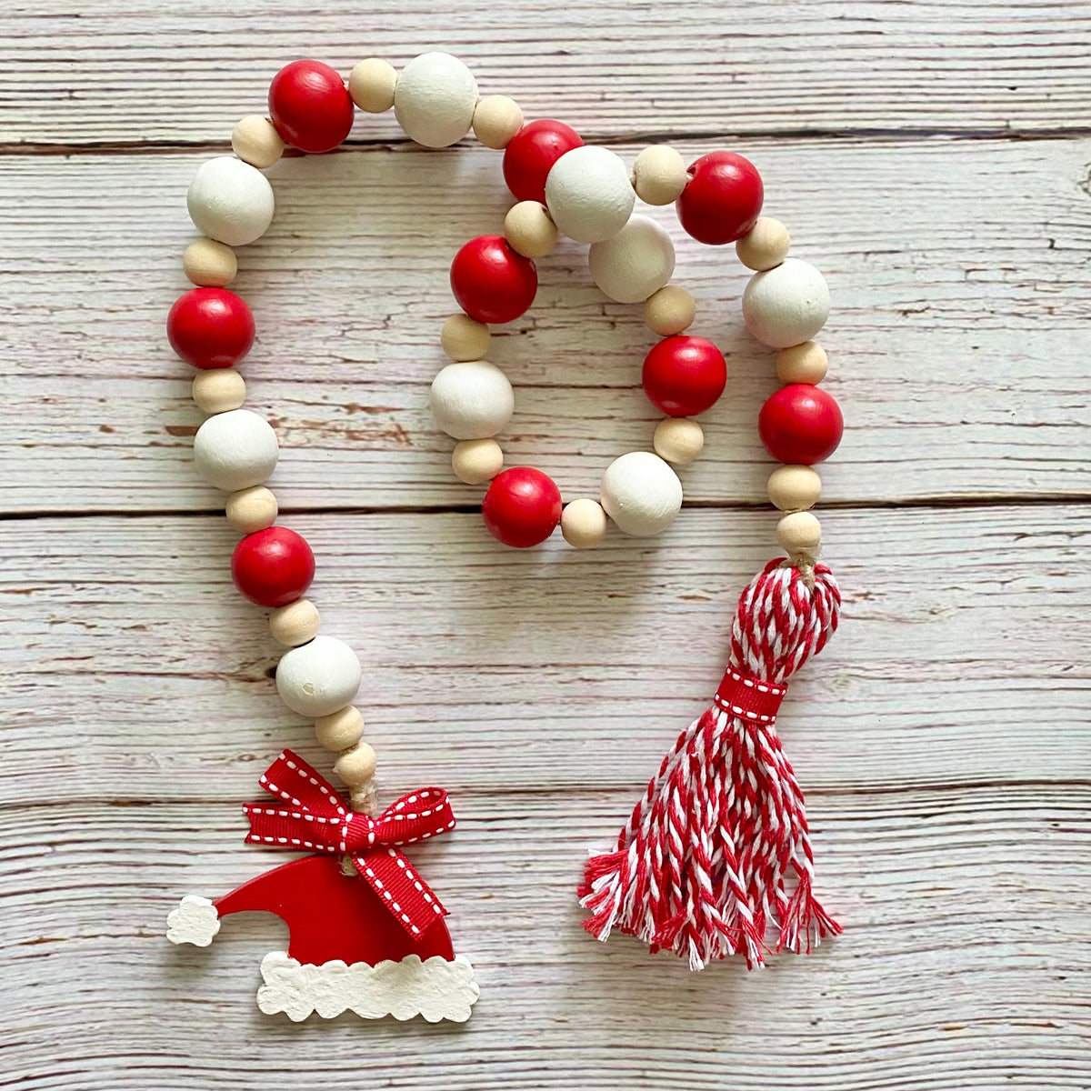 A Christmas-themed wooden bead garland with red and white painted beads, natural wood beads, a red Santa hat ornament, and a red-and-white striped tassel, displayed on a light wood plank background.