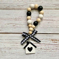 A beaded garland with alternating black and natural wood beads, tied with a black and white gingham ribbon and hanging from a small white birdhouse charm with a black heart, displayed on a rustic whitewashed wooden background.