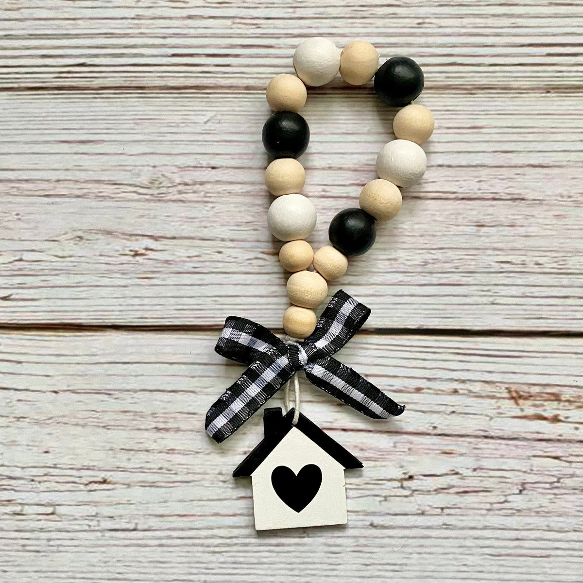 A beaded garland with alternating black and natural wood beads, tied with a black and white gingham ribbon and hanging from a small white birdhouse charm with a black heart, displayed on a rustic whitewashed wooden background.