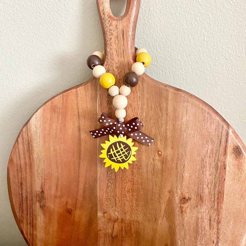 A wooden cutting board with a natural finish, featuring a decorative beaded handle garland with yellow, brown, and cream beads, a brown polka dot ribbon bow, and a yellow sunflower charm.
