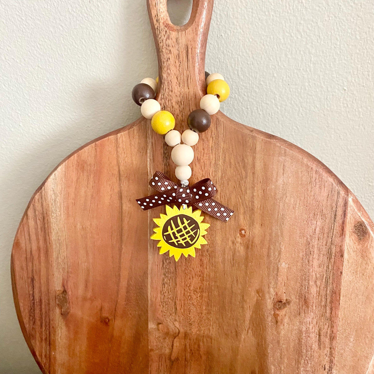 A wooden cutting board with a natural finish, featuring a decorative beaded handle garland with yellow, brown, and cream beads, a brown polka dot ribbon bow, and a yellow sunflower charm.