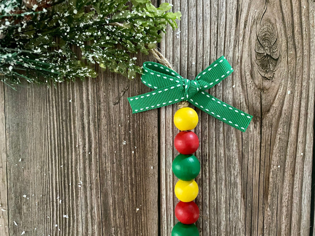 A festive Christmas ornament featuring a green ribbon bow with white stitching, hanging from a string. The ornament's body is made of alternating red, green, and yellow wooden beads, displayed against a rustic brown wood background with a snowy green pine branch in the corner.