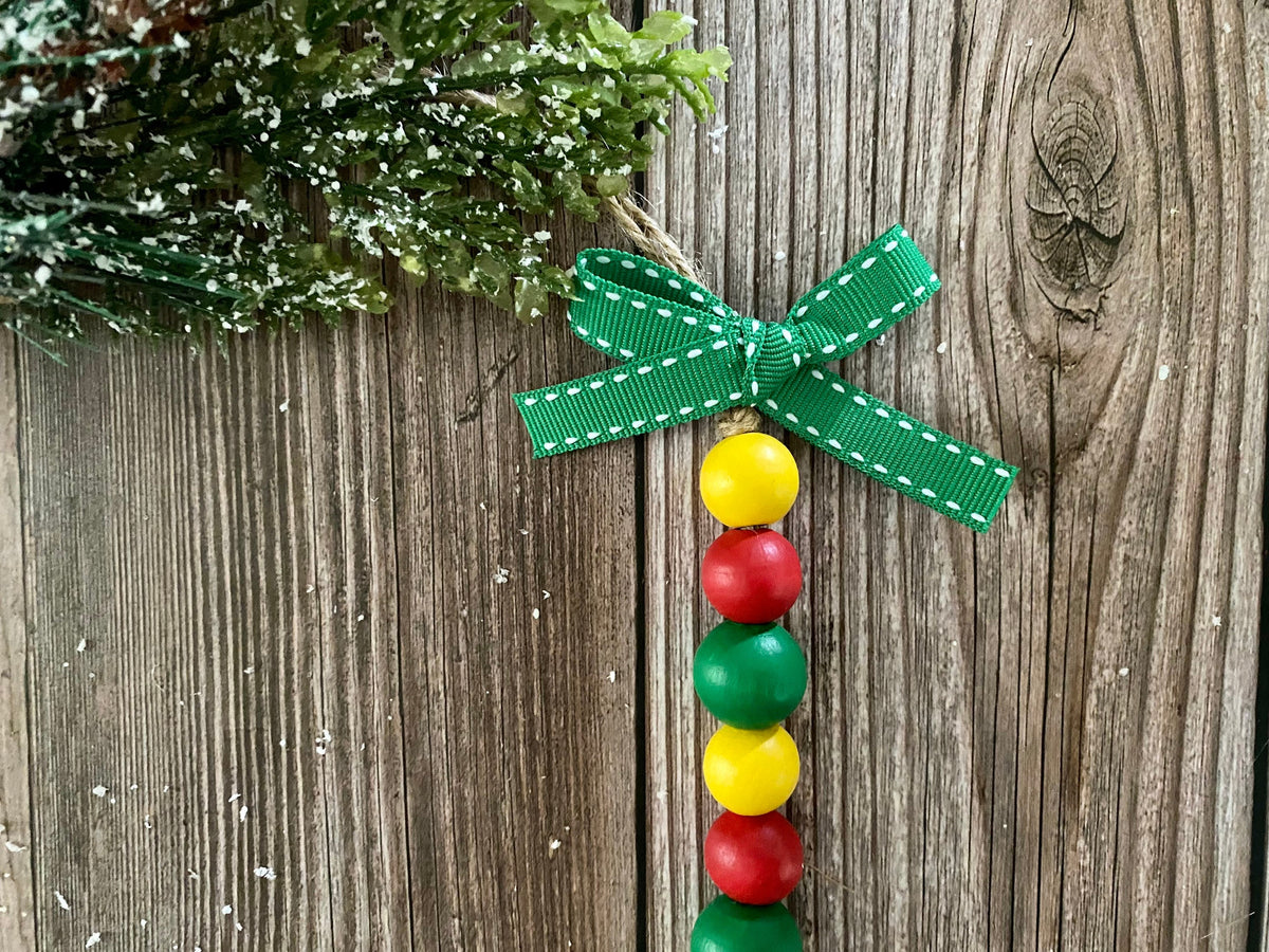 A festive Christmas ornament featuring a green ribbon bow with white stitching, hanging from a string. The ornament's body is made of alternating red, green, and yellow wooden beads, displayed against a rustic brown wood background with a snowy green pine branch in the corner.