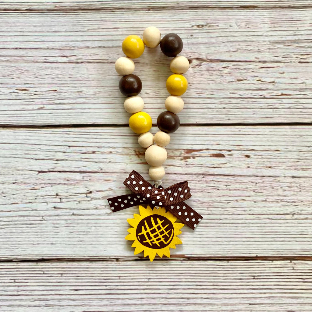 A beaded loop made of alternating yellow, brown, and cream wooden beads, featuring a brown polka-dot ribbon bow and a yellow sunflower charm at the bottom, displayed on a rustic white-washed wooden background.