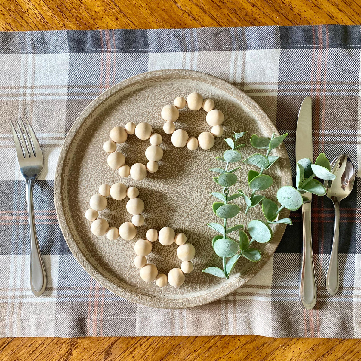 A top-down view of a ceramic plate styled with a beige and brown plaid placemat on a wooden table. The plate features light wood bead garlands and a green eucalyptus sprig, accompanied by silver cutlery.