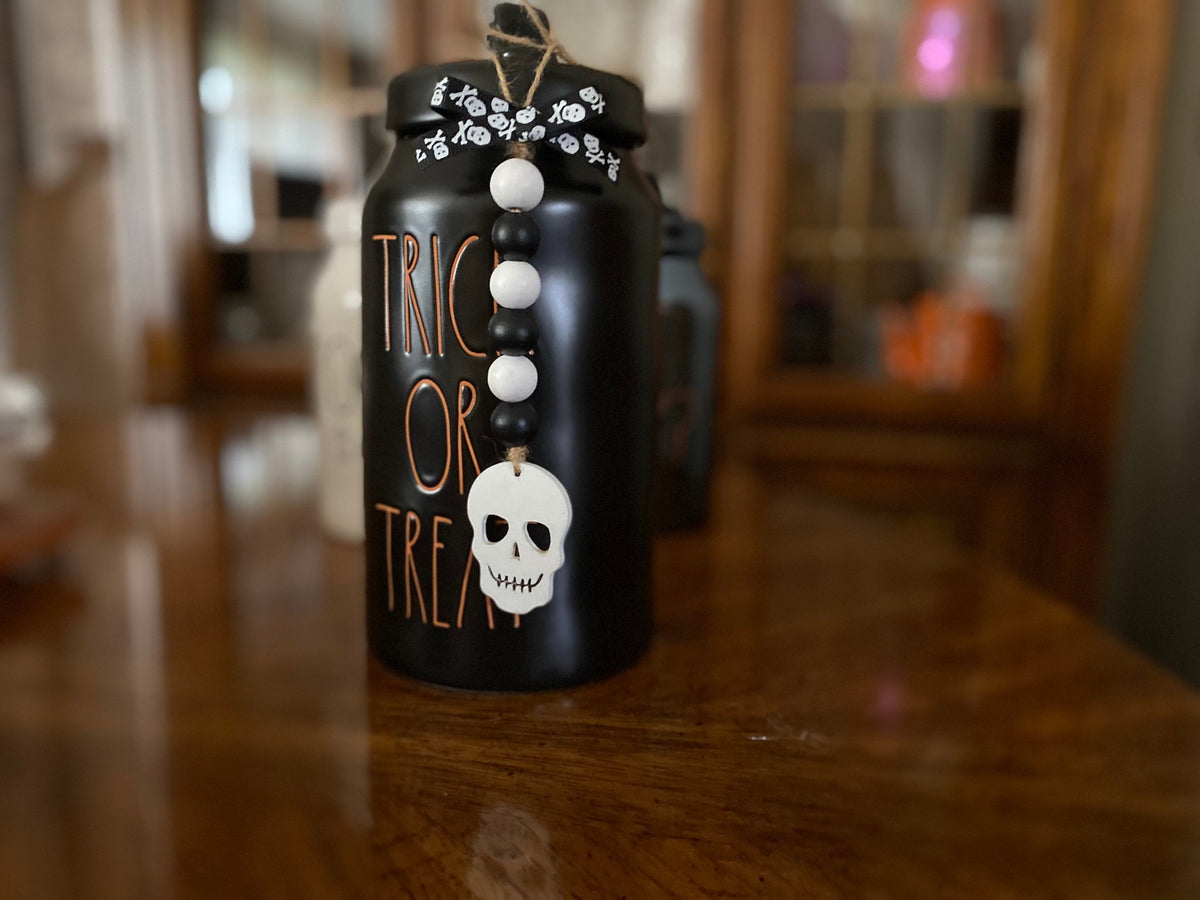 A black Halloween-themed jar or canister with 'TRICK OR TREAT' written in orange lettering, decorated with a white skull charm, white and black beads, and a floral pattern on the lid, sitting on a wooden table.